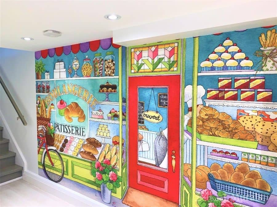 Bakery Wall Mural, as seen in this basement playroom, is a kids wallpaper featuring the front of a bakery with windows looking onto shelves of cookies, cake, pie, candy and other treats from About Murals.