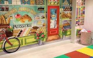 Bakery Wall Mural, as seen in this playroom, features a window full of cakes, cupcakes, croissants and desserts. Kids wallpaper sold by AboutMurals.ca.