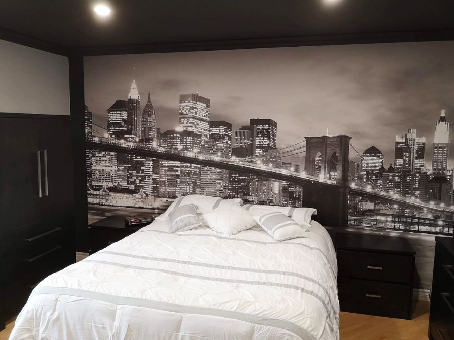 NYC Bridge Wallpaper, as seen in this dark bedroom with a white bed, is a photo wallpaper of the famous bridge in New York City against skyscrapers from About Murals.