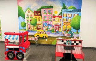 Candy Street Wall Mural, as seen in this Indoor Playground called Batter Zone in Hamilton, Ontario, is a kids wallpaper featuring a cute city with a bakery, chocolate store, pet shop, circus school and toy shop from About Murals.