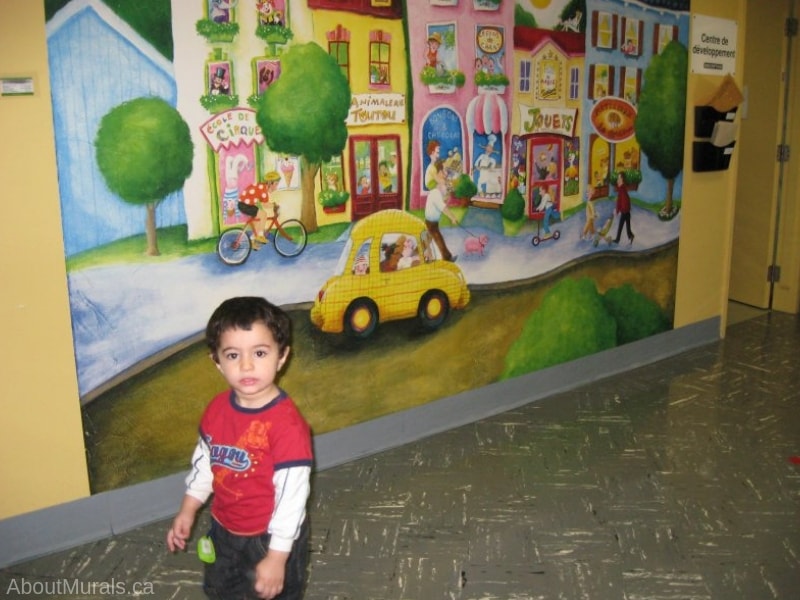 Children’s Town Wallpaper Mural, as seen on the wall of a doctor office, is an uplifting kids wall mural with people walking past a colourful circus school, pet shop, candy store, toy shop and bakery from About Murals.