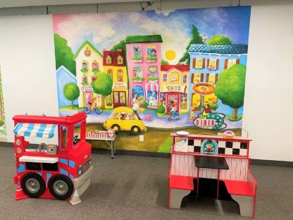 Children’s Town Wallpaper Mural, as seen on the wall of this preschool, inspires hours of imaginative play in a cartoon town with people walking past a circus school, pet shop, candy store, toy shop and bakery from About Murals.