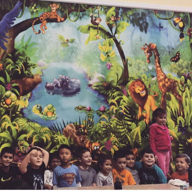 Jungle Wall Mural, as seen behind eleven children in this jungle safari indoor playground, features animals in a lush rainforest near a blue pond from About Murals.