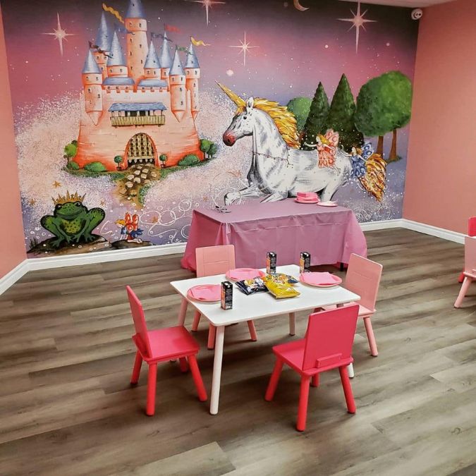 Princess Castle Wallpaper, as seen on the wall of this preschool, is a toddler mural with a pink castle, white unicorn and green frog prince on a purple background from About Murals.