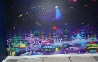 Space City Wall Mural, as seen at this indoor playground, is a kids space wallpaper with UFOs flying over a purple intergalactic city from About Murals.