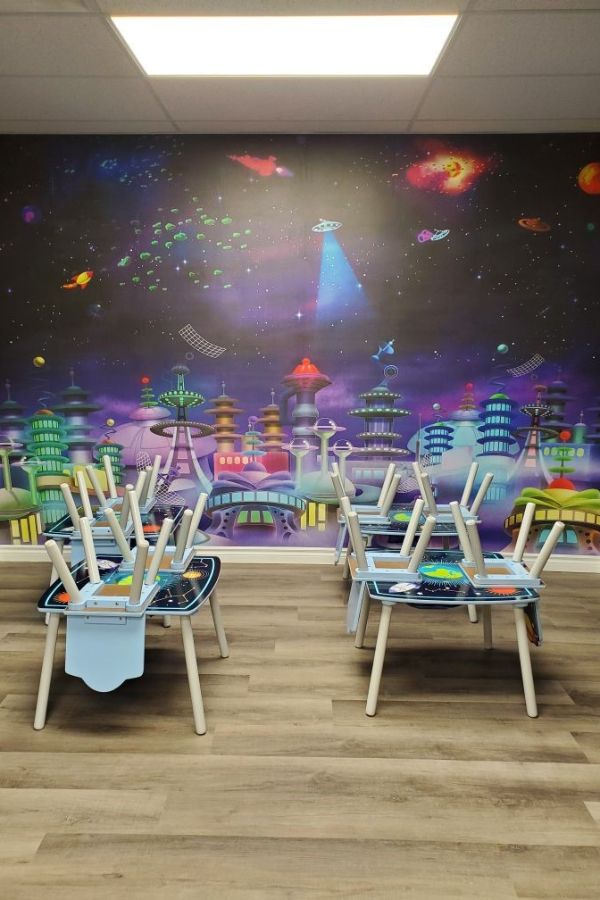 Space City Wall Mural, as seen in this preschool, is a space themed wallpaper with spaceships flying over skyscrapers and buildings in a purple galaxy from About Murals.