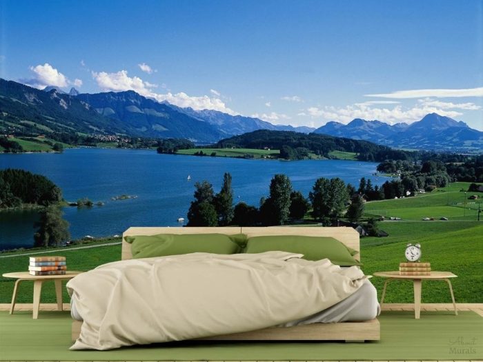 Switzerland Wall Mural, as seen in this bedroom, features blue mountains towering over a lake and green grass. Landscape wallpaper sold by About Murals.