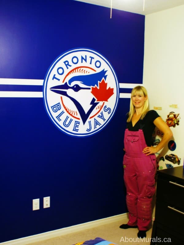 Muralist, Adrienne of AboutMurals.ca, stands next to a baseball mural she painted of a Toronto Blue Jays logo