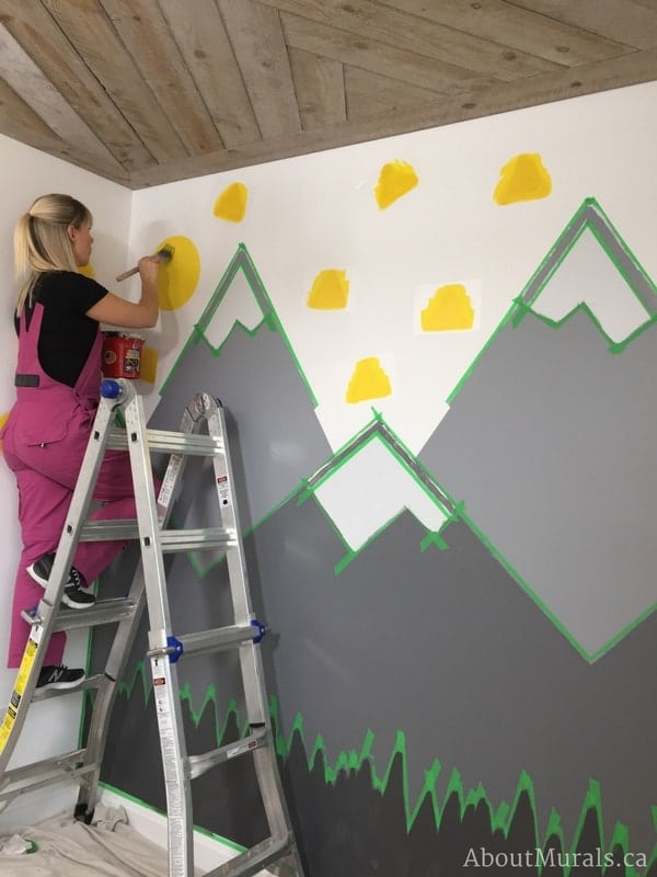 Holmes Next Generation contractor, Adrienne of AboutMurals.ca, paints a yellow sun and triangles in the mountain mural