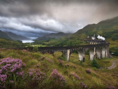 Steam Train Wallpaper is a photo mural of an old train chugging over an arched bridge in Scotland under stormy clouds that settle over mountain tops from About Murals.
