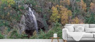 Autumn Waterfall Wallpaper, as seen on the wall of this living room, is a photo mural of small falls spilling over a large rock face surrounded by green and orange trees in a forest from About Murals.