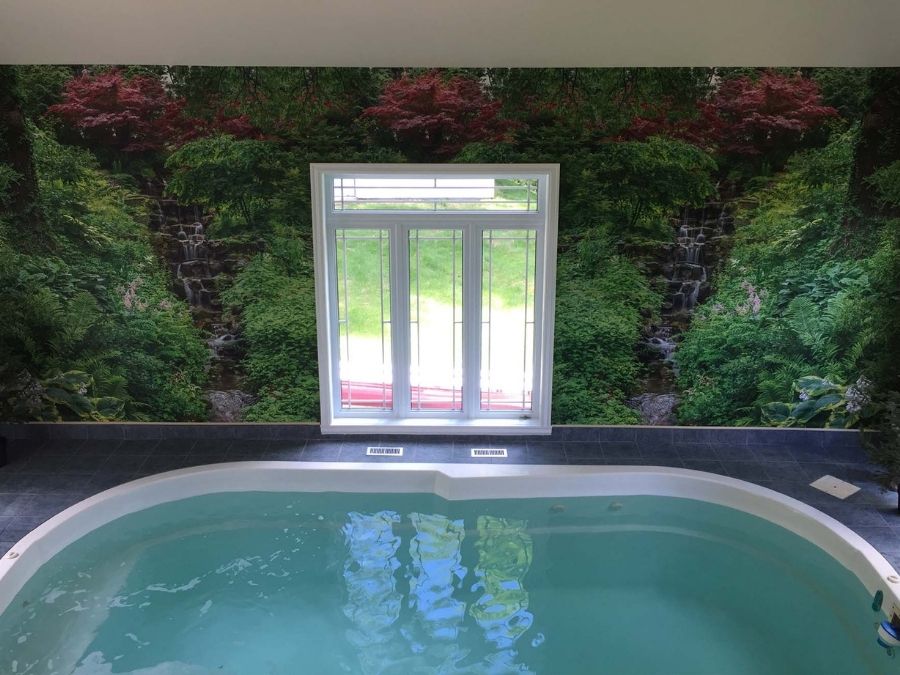 Green Waterfall Wallpaper, as seen in this indoor pool, is a photo mural of water gushing down brown stone in a park garden from About Murals.