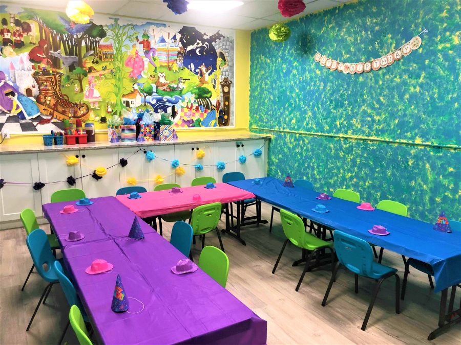 A fairytale themed birthday party venue wallpaper mural at an indoor playground behind a party table, children’s plastic chairs, banners and party hats.