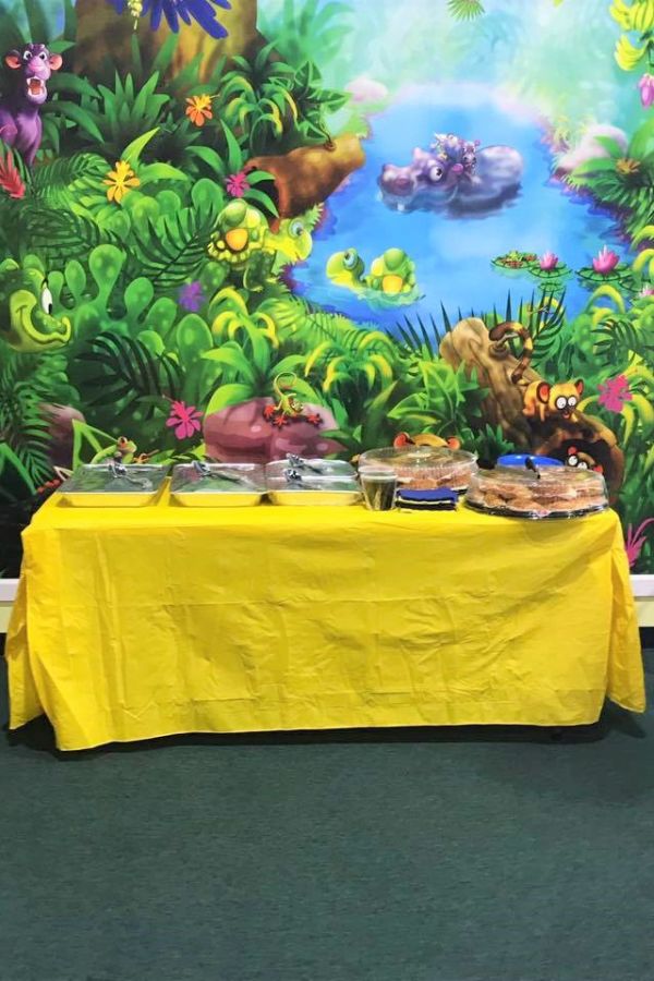 Jungle party room wallpaper mural, featuring animals like a monkey, panther, hippo, turtle and snake, on the wall behind a birthday party table with food.